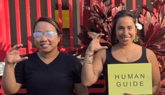 Two women demonstrating human guide techniques during an immersive workshop on blindness awareness. One participant is wearing vision simulation glasses, while the other holds a sign that reads 'HUMAN GUIDE,' highlighting training in sighted guide techniques.