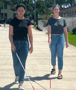 Two young women participating in a vision loss awareness lesson. One walks with her eyes closed, simulating blindness, while the other uses a white cane, practicing independent mobility skills in an outdoor setting.