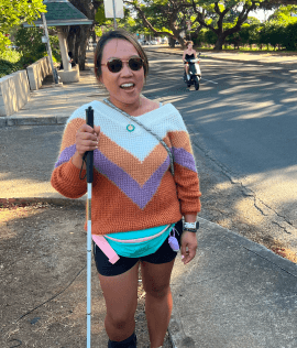 A woman with a white cane participating in a vision loss awareness lesson outdoors. She is wearing sunglasses and a colorful sweater, confidently navigating the environment, promoting blindness awareness and mobility skills.