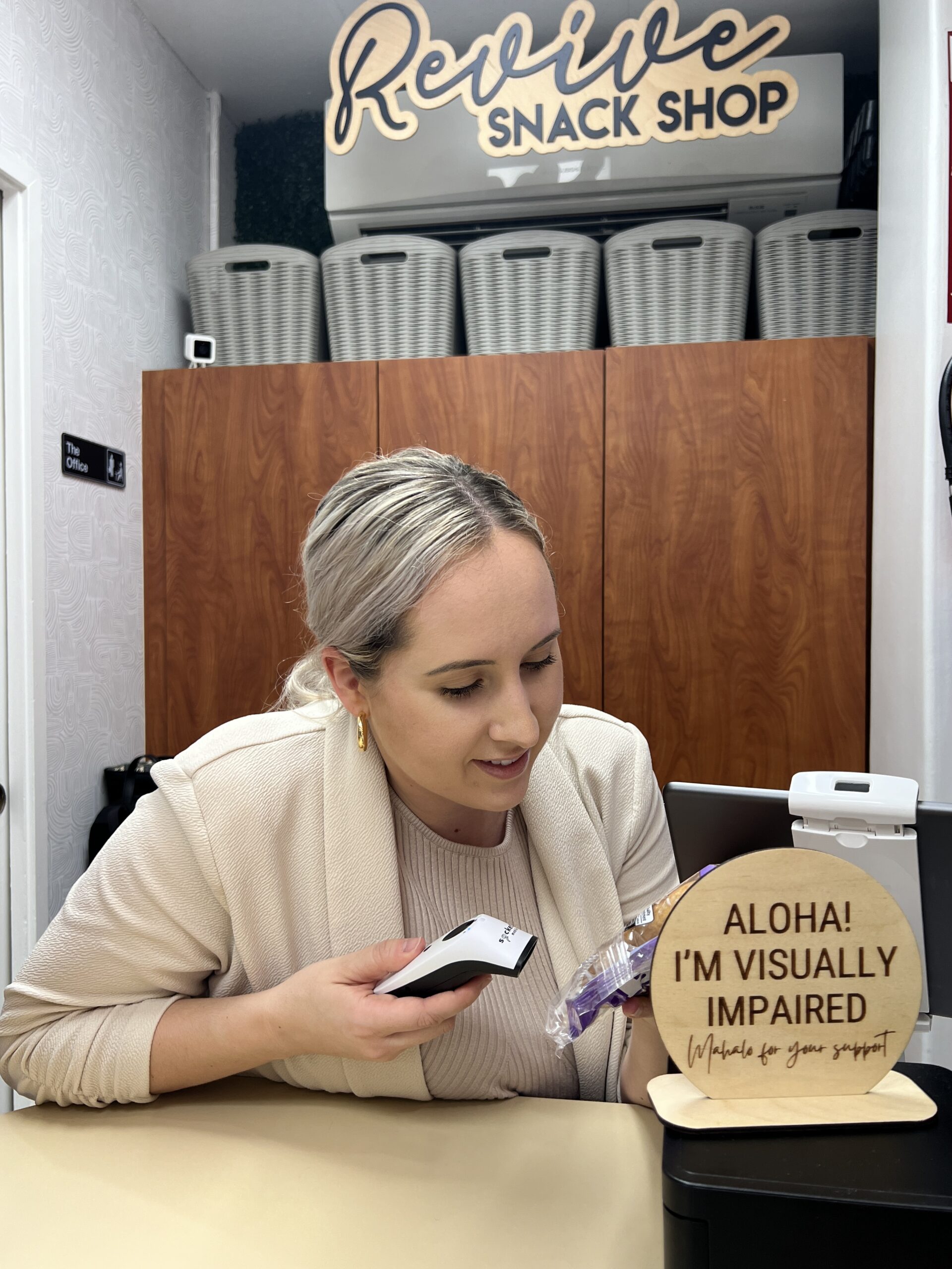 Woman working in a snack sho leaning close to scan a product with an engraved sign that reads “Aloha, I’m Visually Impaired, Mahalo for your Support”