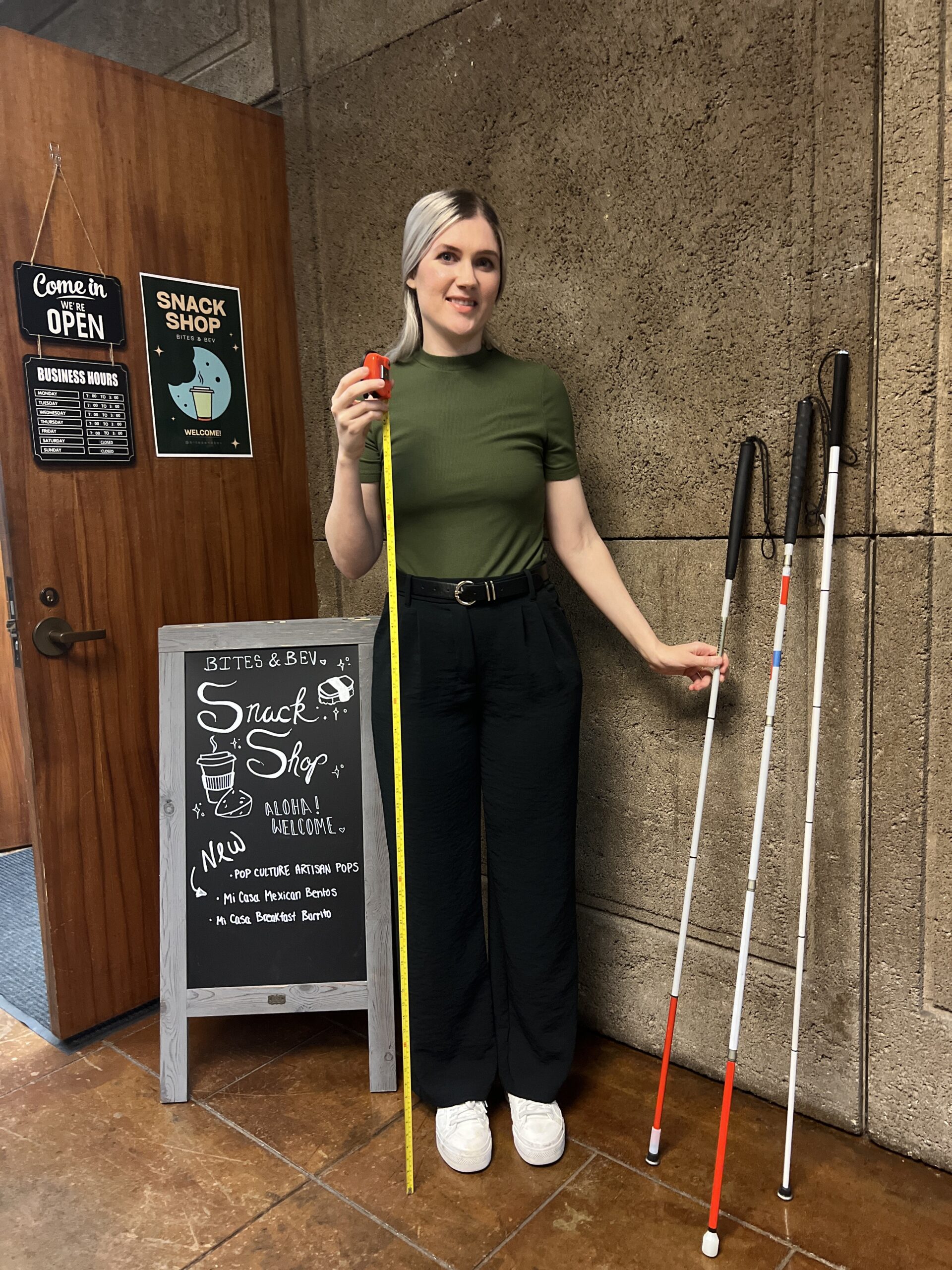 Woman outside of a snack shop measuring her cane size with three white canes to her left.